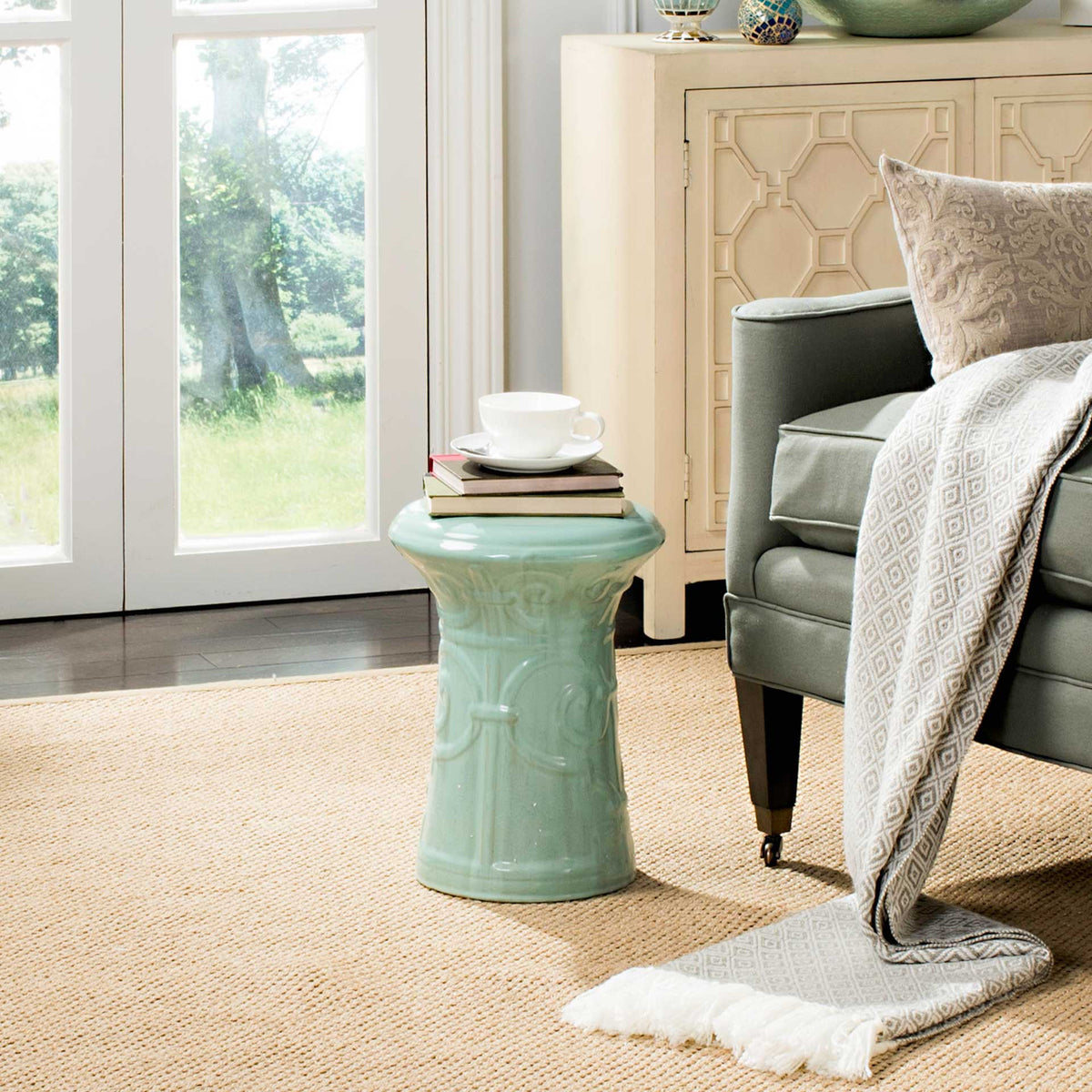 A light aqua ceramic garden stool holds stacked books and a cup beside an upholstered chair