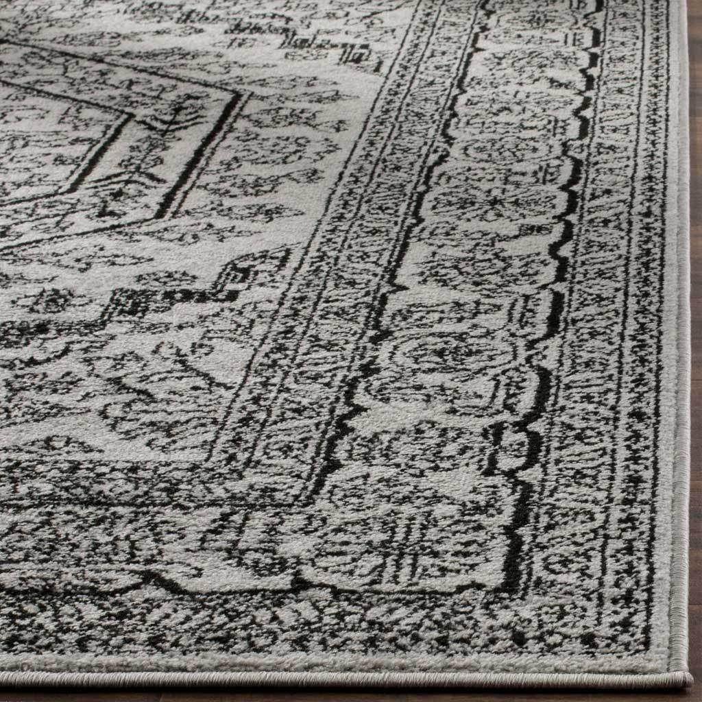 Black floral borders ripple across soft silver pile to a carefully bound rug corner.