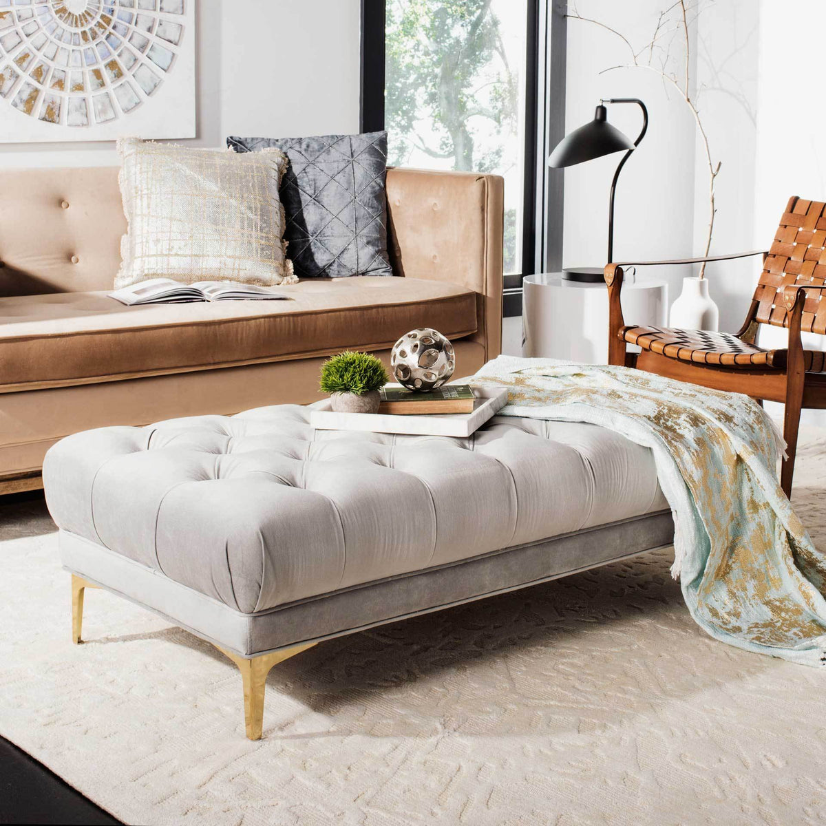A plush gray velvet bench doubles as a coffee table, with deep tufting and tapered brass-finished legs.