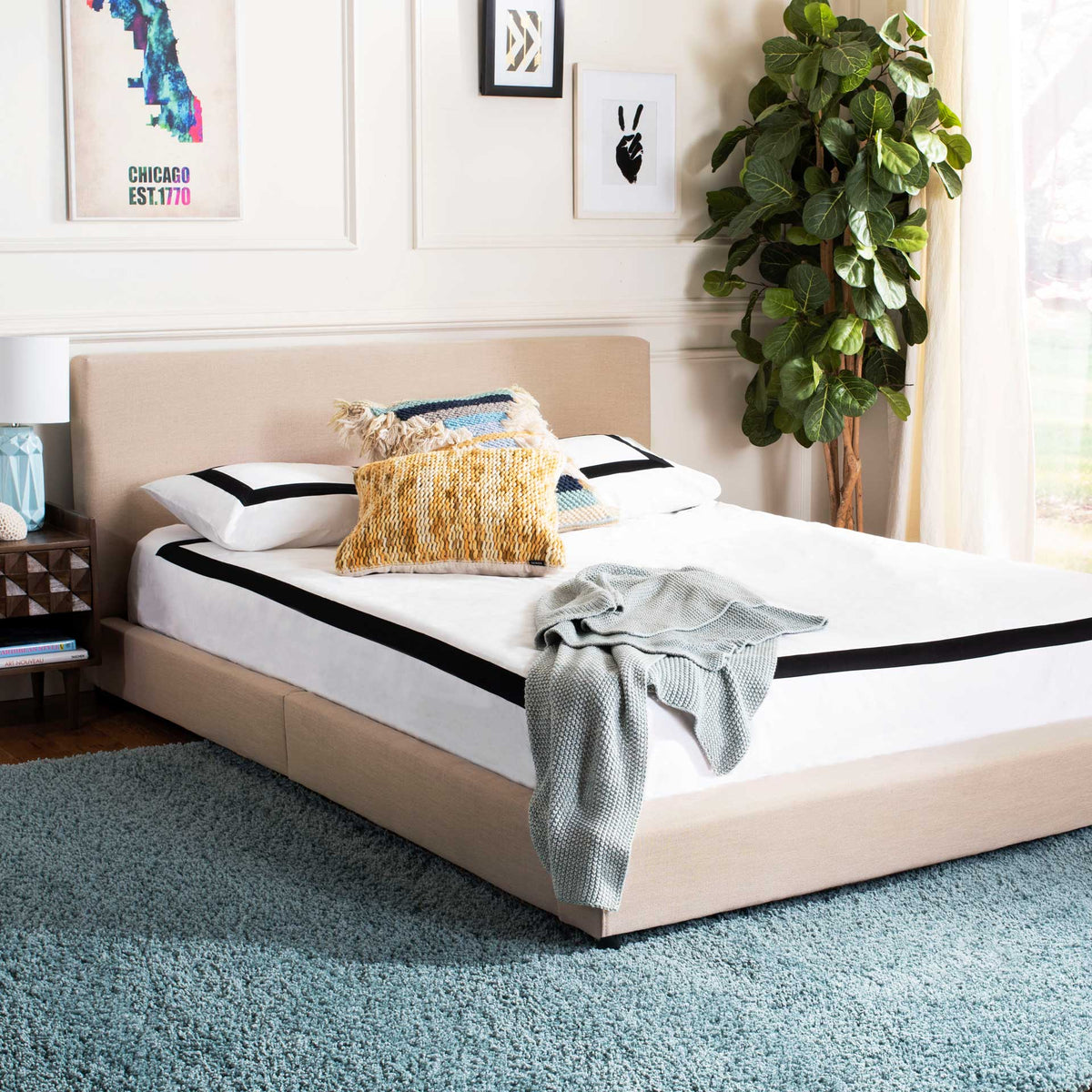 The beige linen bed has a broad, cushioned headboard and a low-profile upholstered frame