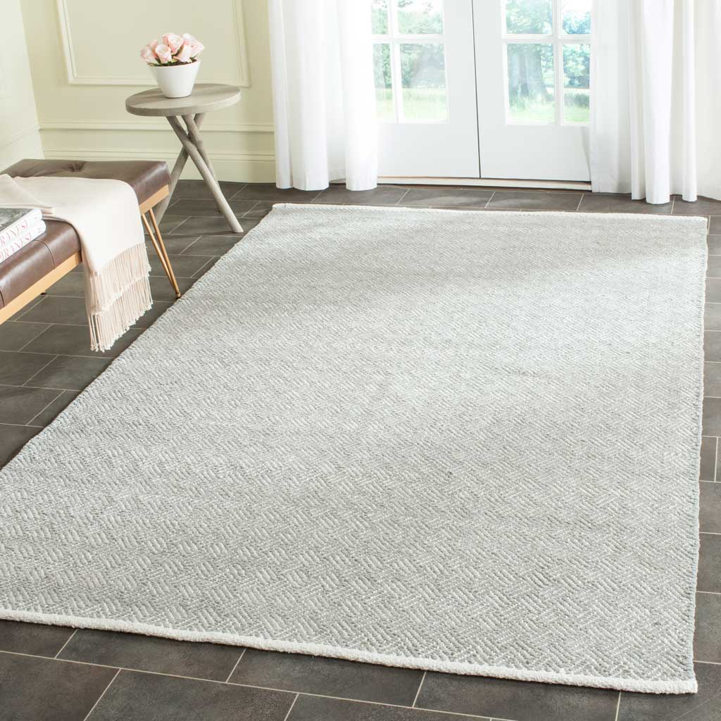 A light gray herringbone rug brightens dark tile with soft cotton texture and ivory edging.