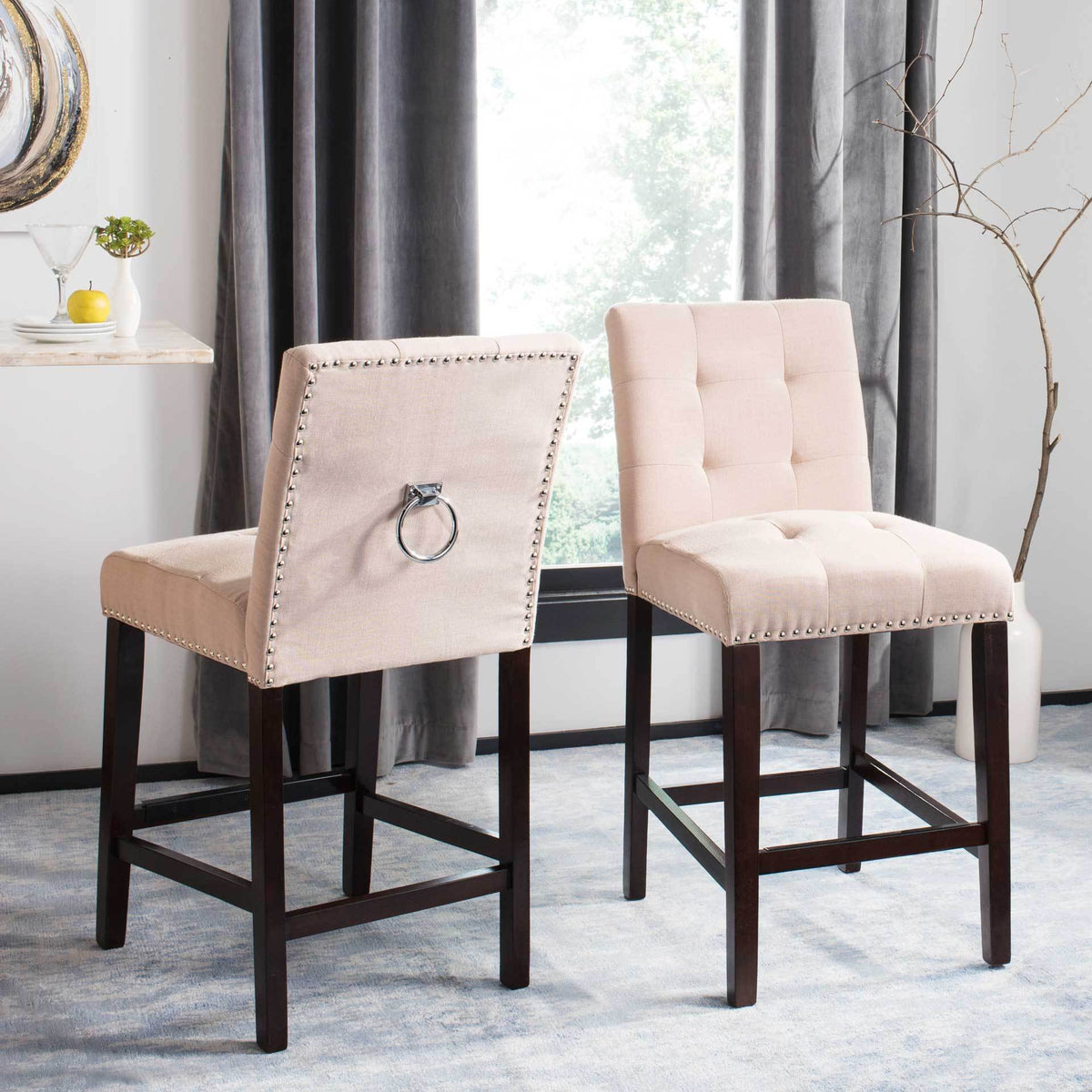 Two beige counter stools pair tufted linen seating with studded backs and chrome ring details