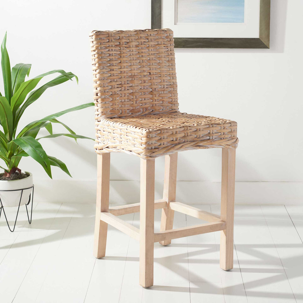 Light wicker counter stool adds a coastal touch beside leafy greenery and a framed print