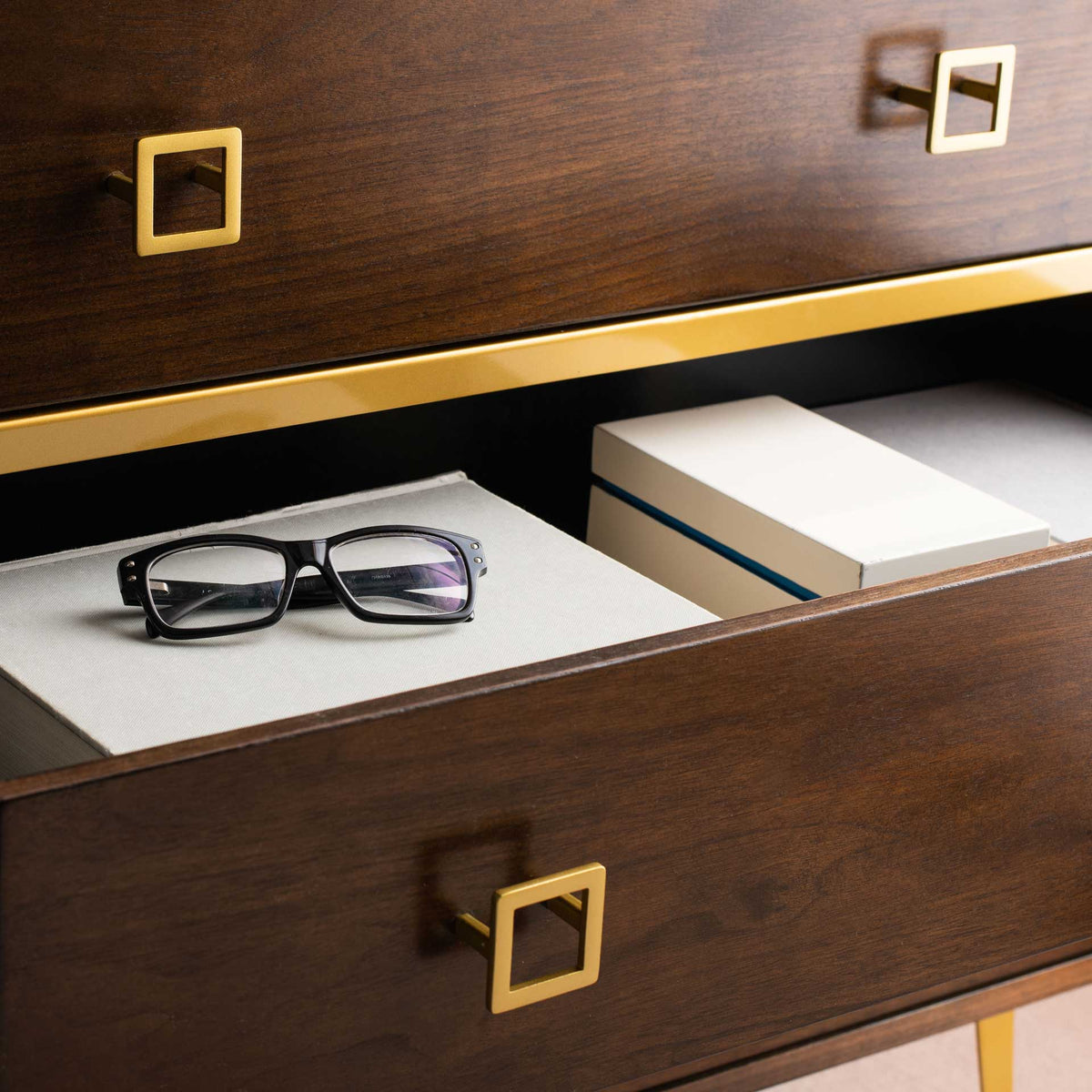 An open walnut drawer holds books and glasses below polished gold accents.