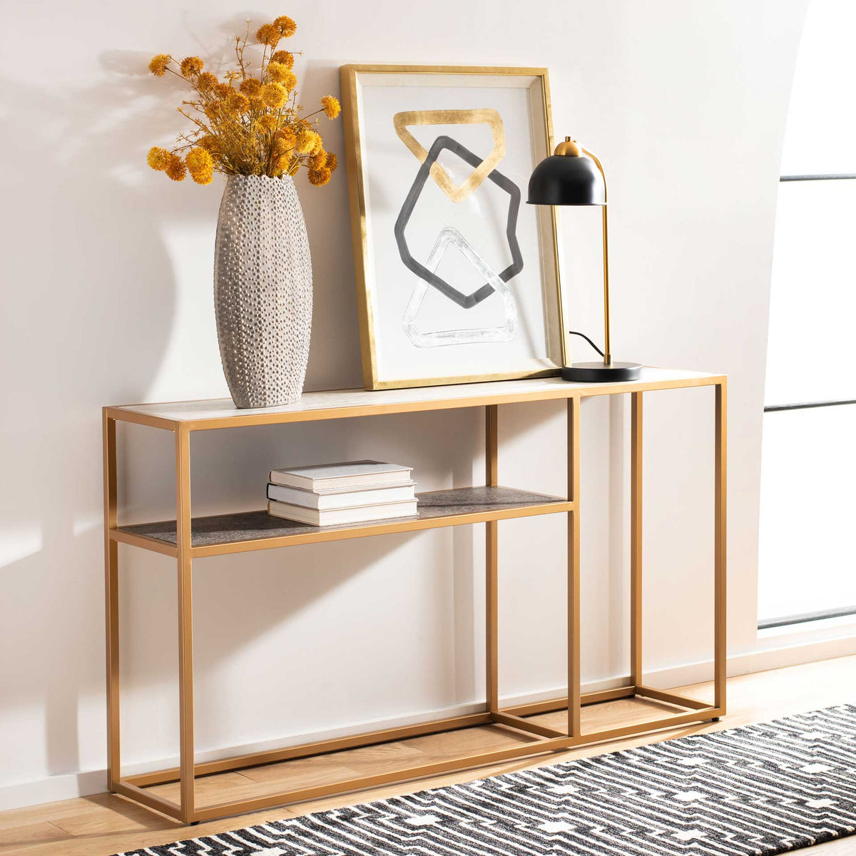 A slim gold console pairs a pale top with a dark shelf, styled with flowers, books, and art.