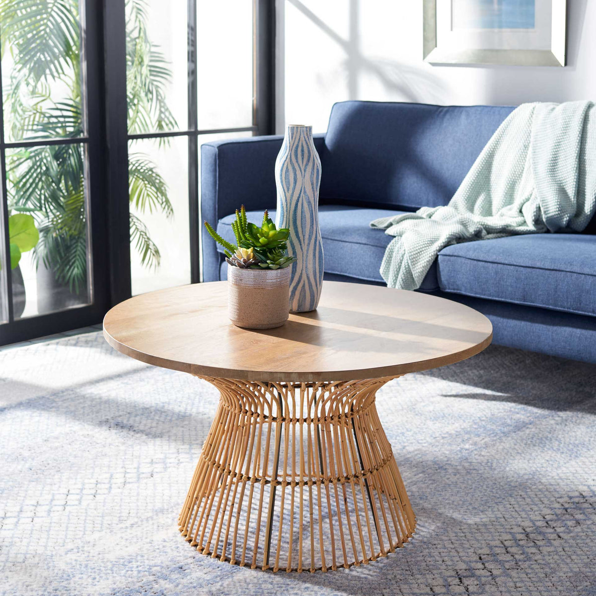 A light wood tabletop and golden rattan pedestal add warm, airy texture beside a blue sofa.