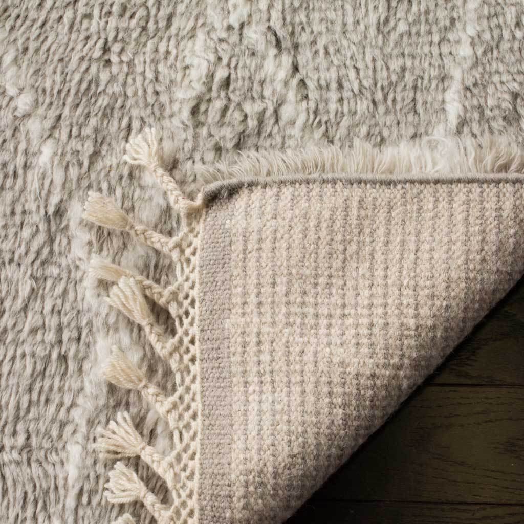A turned edge reveals woven wool backing, thick gray pile, and twisted fringe