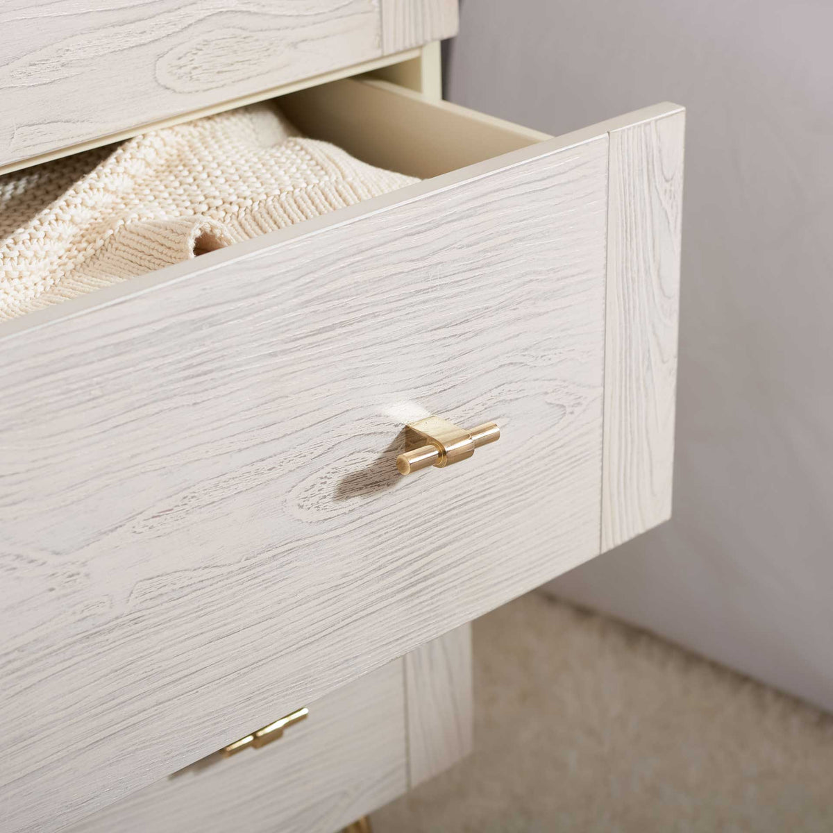 An open drawer holds folded knitwear, showing off whitewashed grain and a gleaming gold pull.