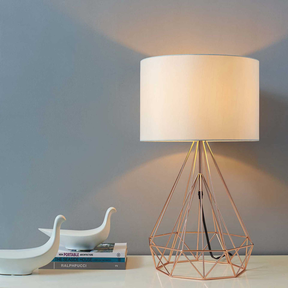 Warm light glows through a cream drum shade above the sculptural rose gold wire base.