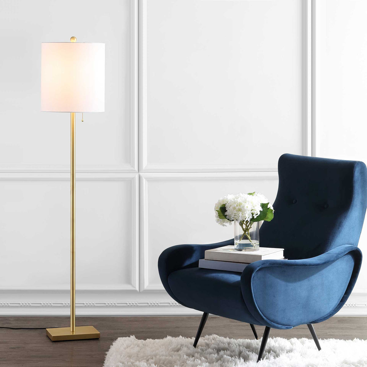 A glowing off-white drum shade brings warm light beside a blue chair on a slim brass-finished floor lamp.