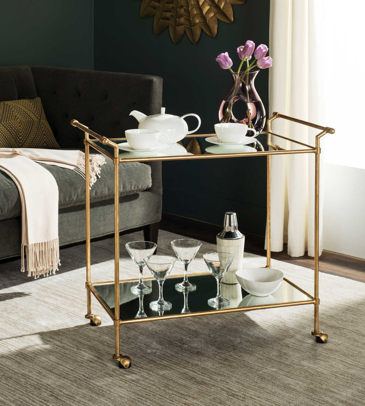 Gold bar cart holds tea service above and glassware below on mirrored shelves