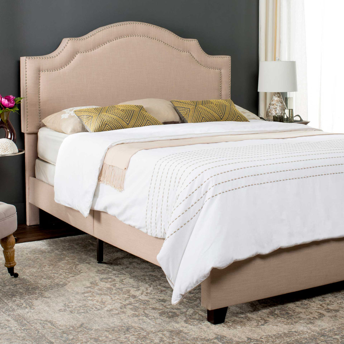 Curved light beige headboard with silver nailhead trim rises above an upholstered bed frame