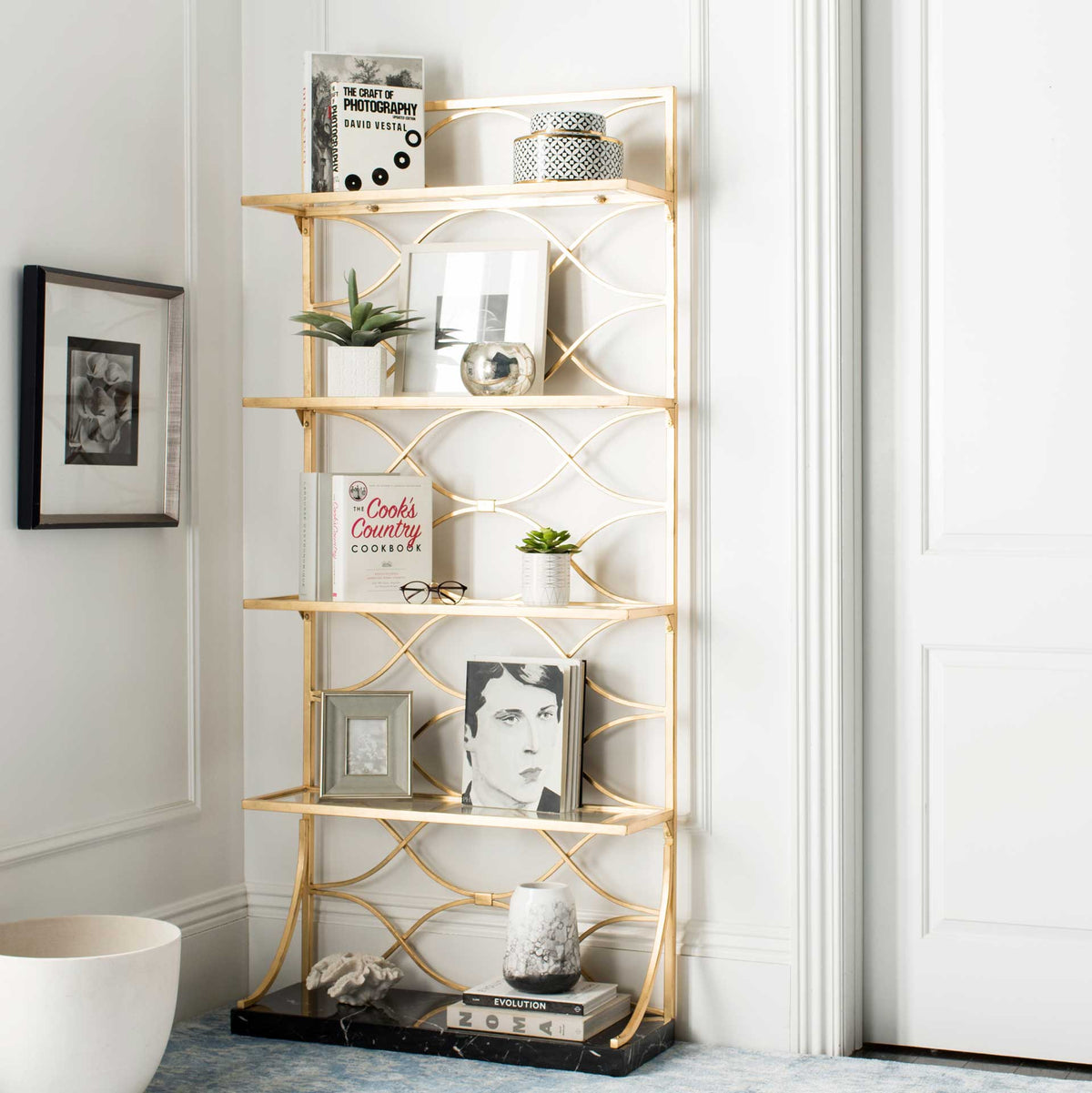 Four glass shelves display books, plants, and decor in an airy gold frame above a black marble veneer base.