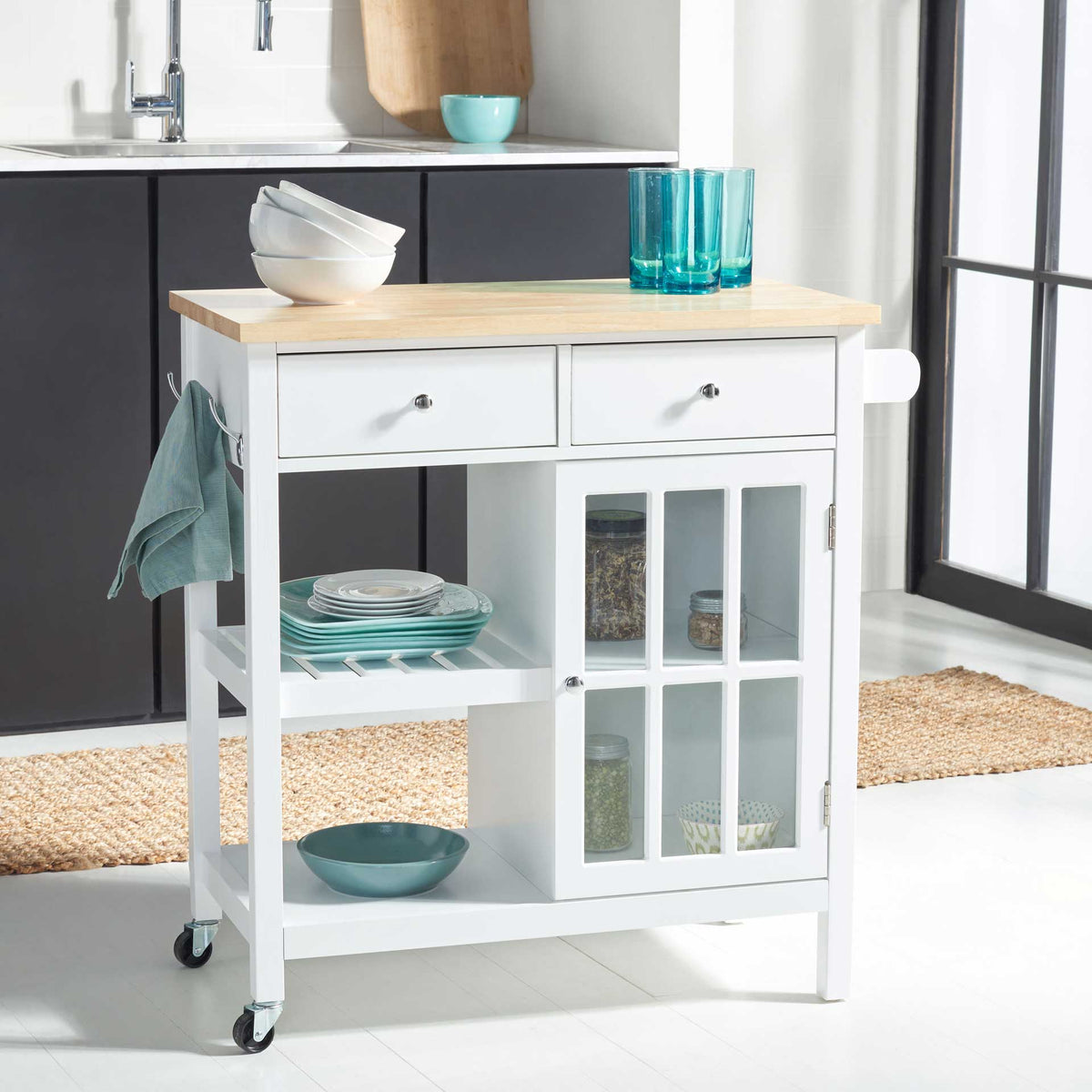 A white kitchen cart stores stacked dishes on open shelves and jars behind glass beneath a natural wood top