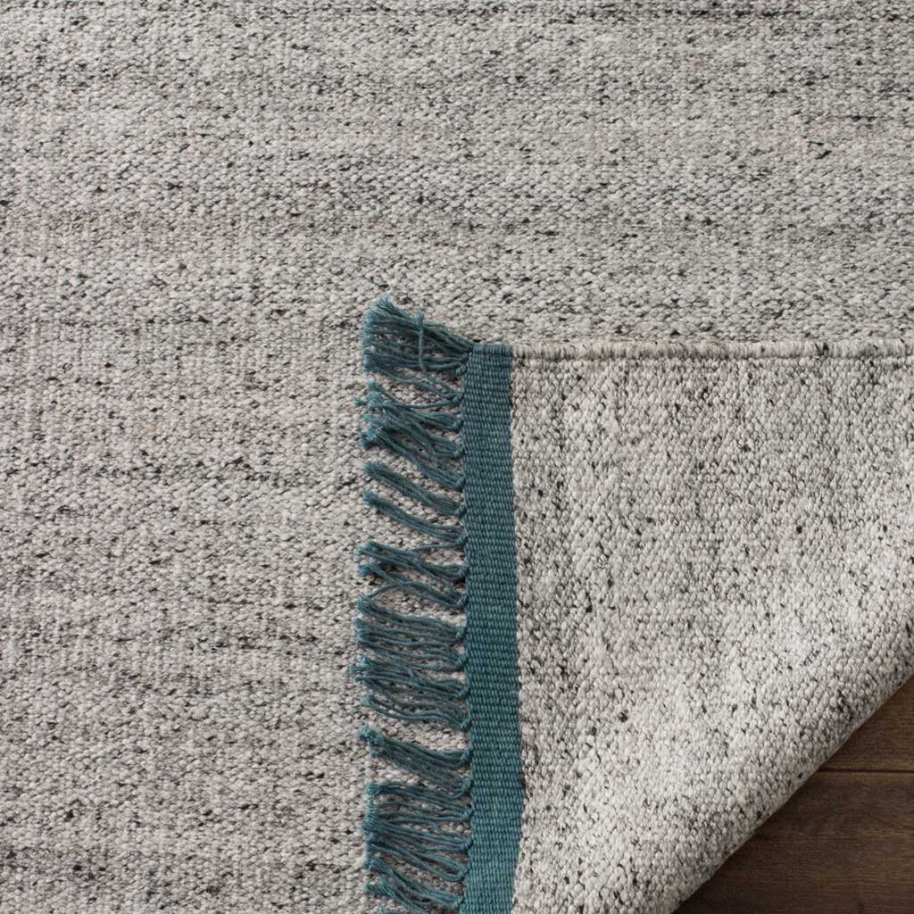A folded corner reveals the rug's woven underside beside teal binding and fringe