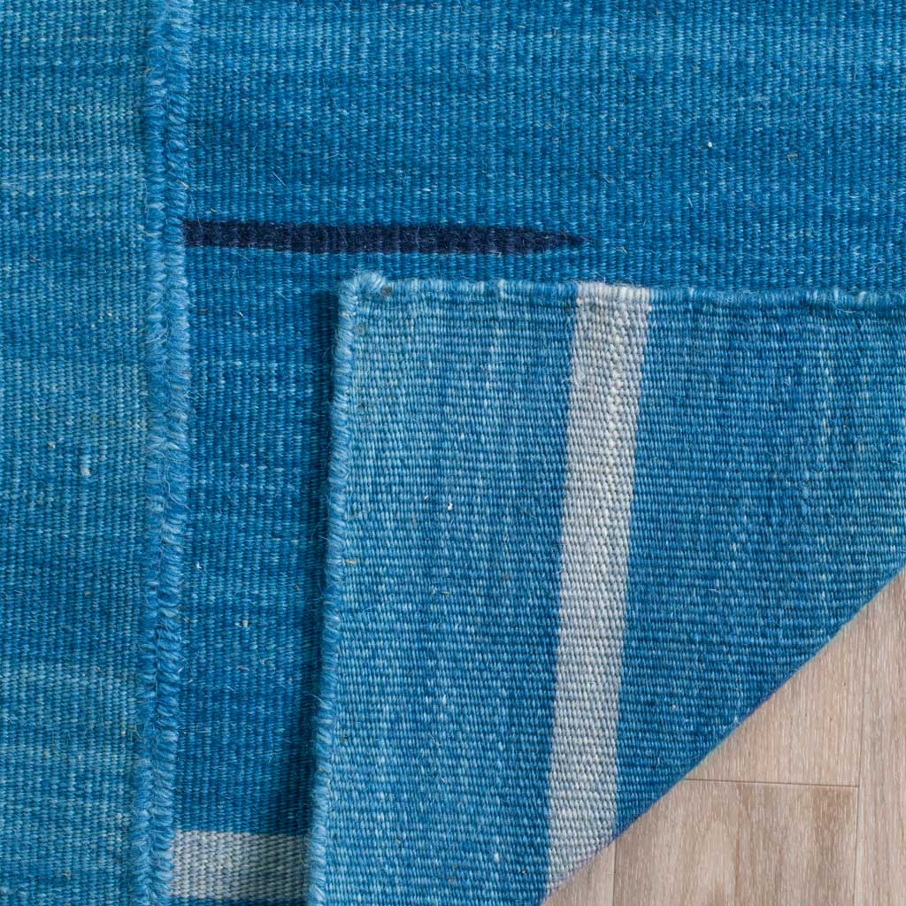 Folded rug edge reveals blue woven backing, a pale stripe, and neat edge stitching.