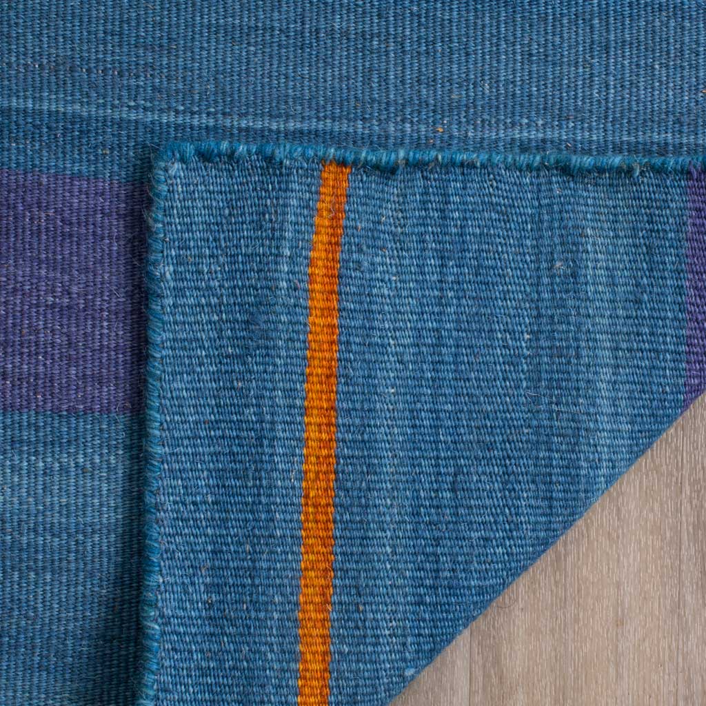 A folded corner reveals the rug's woven reverse, orange striping, and finished blue edge