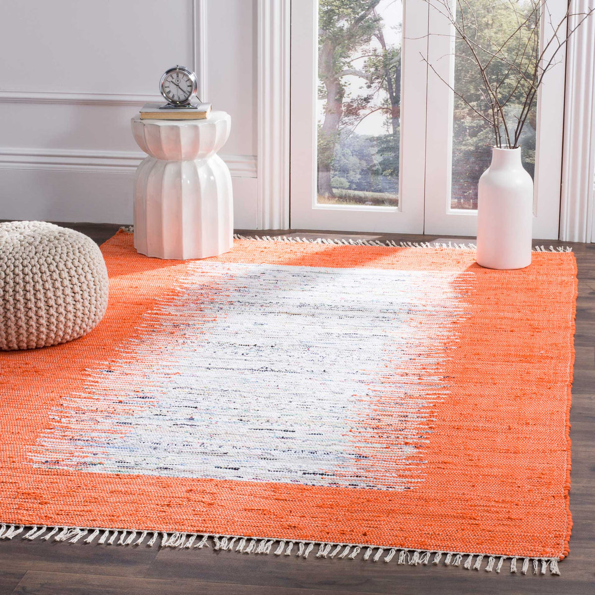 A vivid orange and ivory rug brightens a sunlit seating space with playful woven texture and pale fringe.