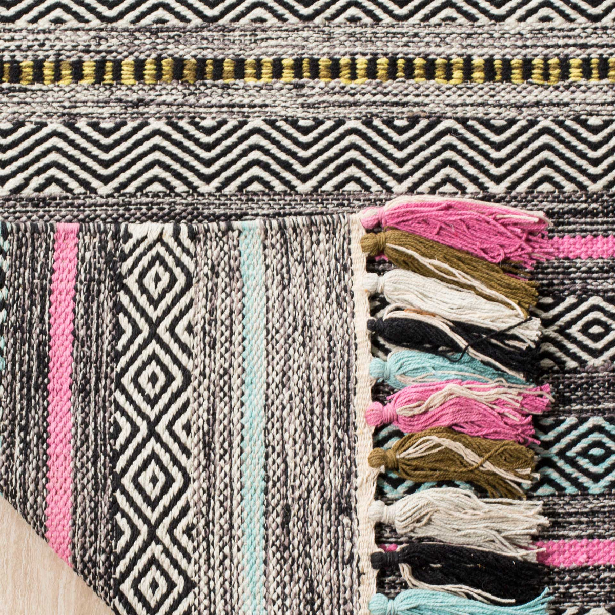 Folded rug edge reveals a striped cotton underside and layered pink, ivory, and aqua tassels.