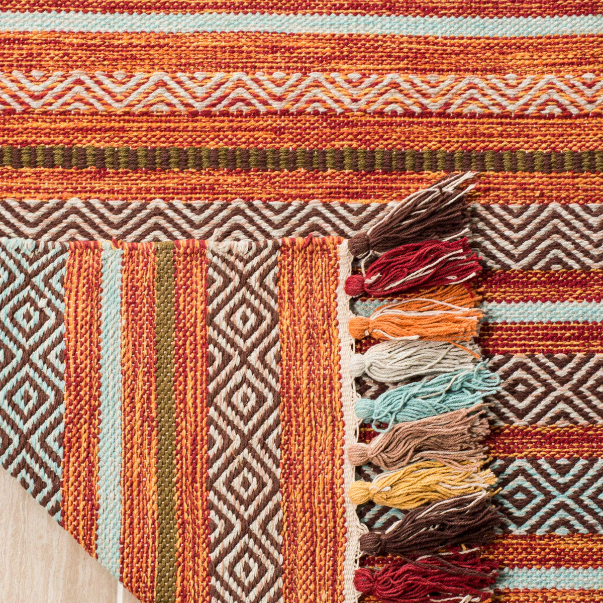 Folded woven corner reveals orange and aqua stripes underneath a vivid tasseled border.