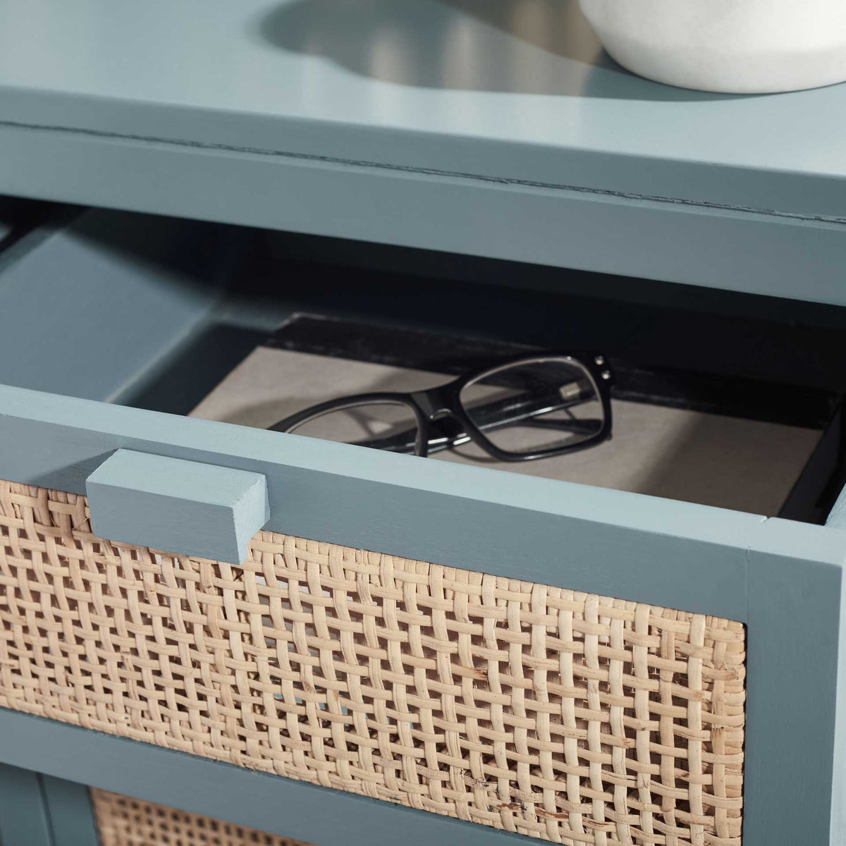 An open blue-gray drawer holds eyeglasses behind its woven cane front.