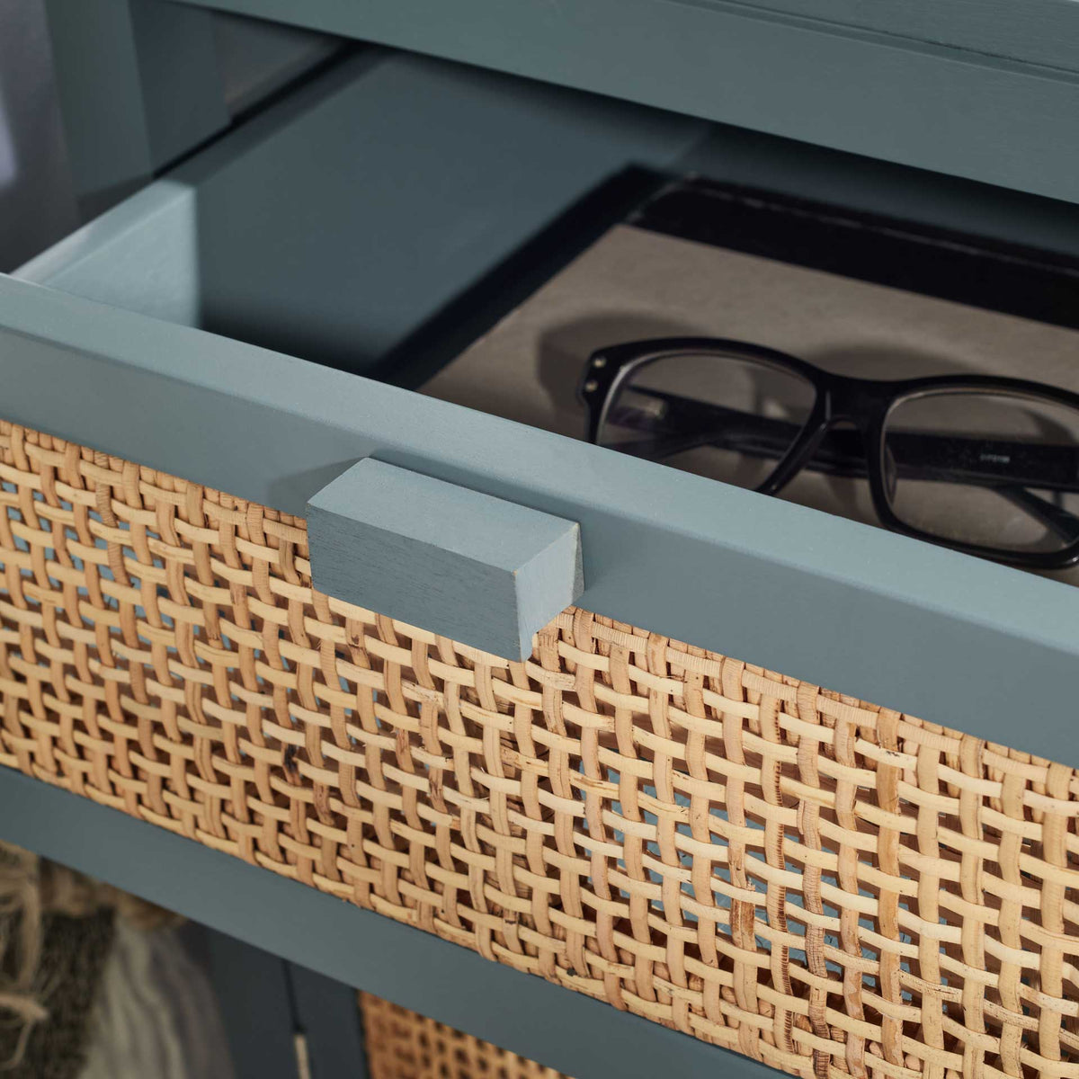 Open cane-front drawer holds eyeglasses in a blue-gray bedside cabinet