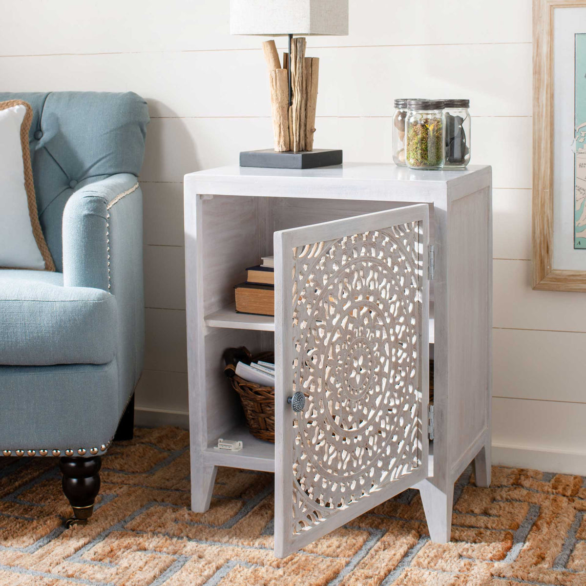 An open carved door reveals divided storage for books and baskets inside the whitewashed nightstand