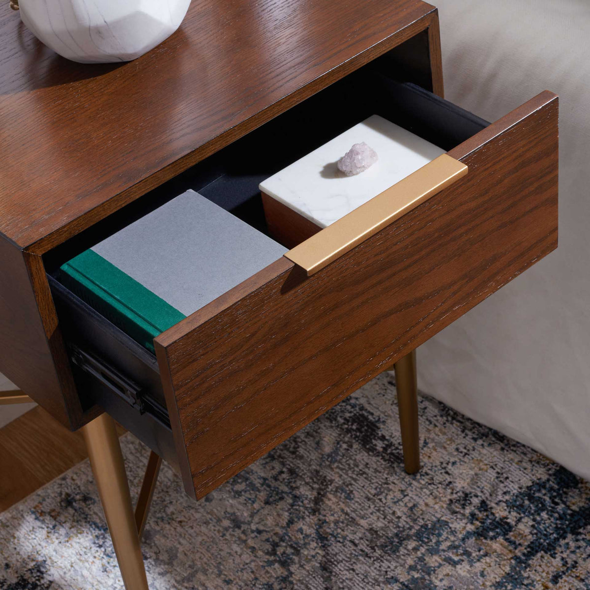 A black-lined drawer holds bedside essentials beneath a walnut-grain top and sleek gold pull.