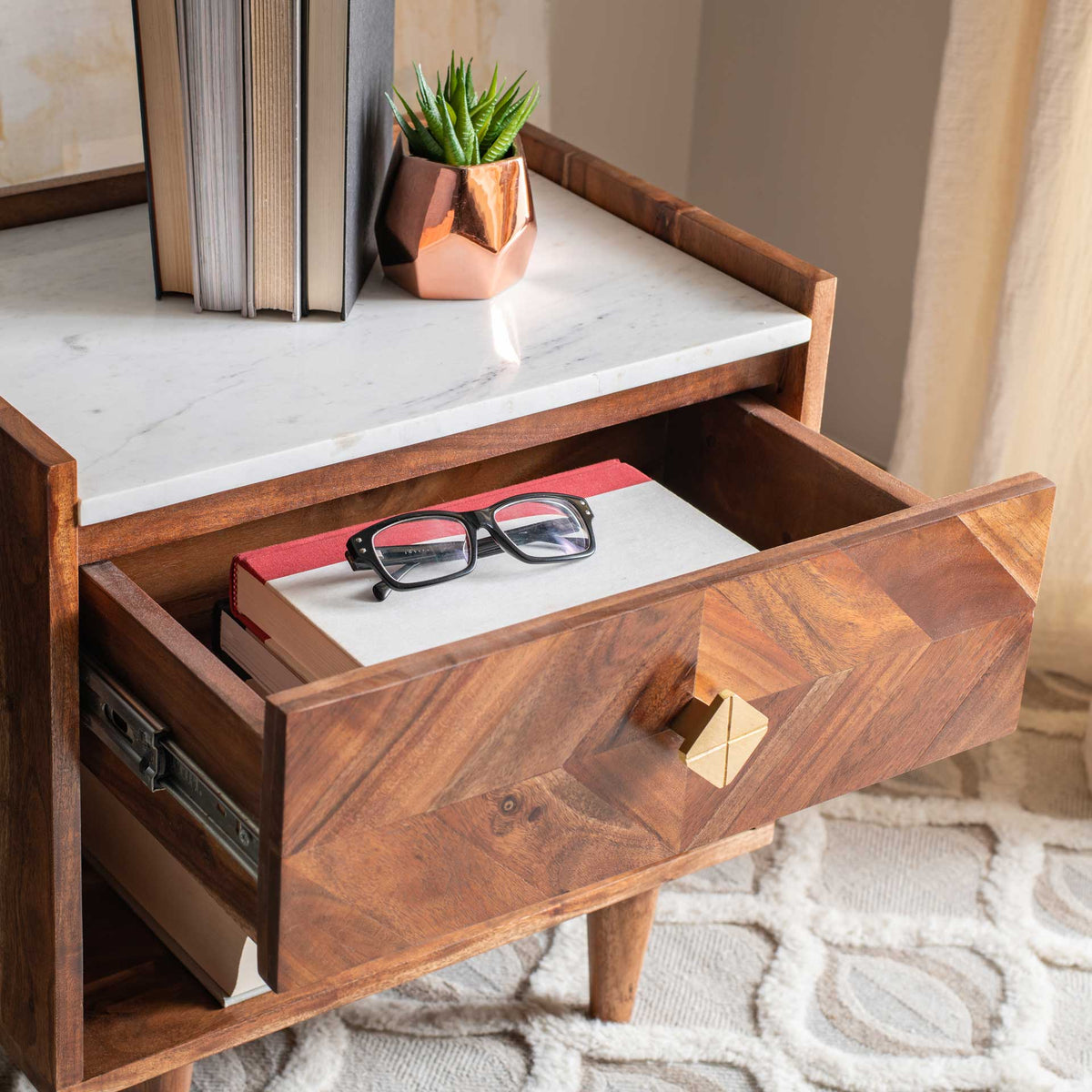 A pulled-out wood drawer stows books and eyeglasses beneath a marble-look top and geometric gold-toned knob.