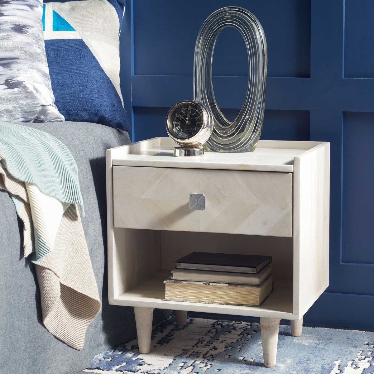 A silver-toned knob brightens the whitewashed parquet drawer over an open bedside shelf and tapered legs.