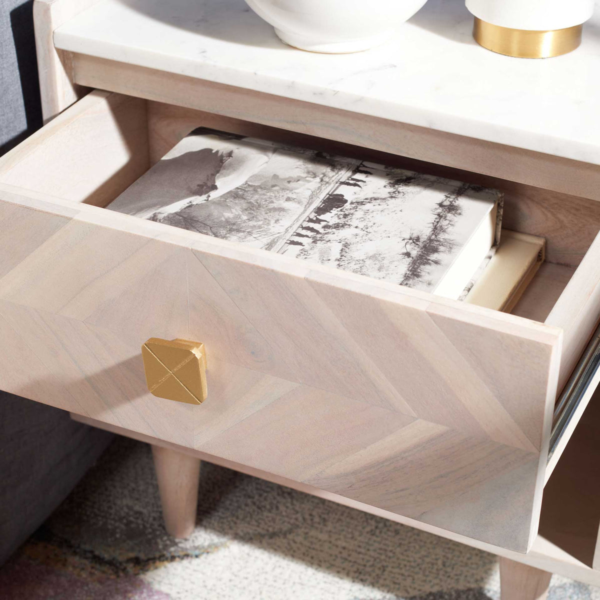 A whitewashed drawer holds books below the marble-look top, with a geometric brass-toned knob up front.