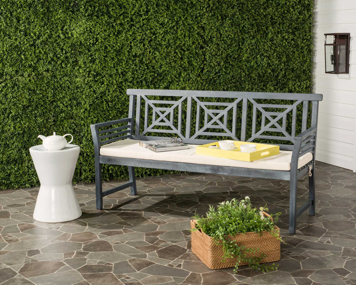 An ash gray wood bench brings geometric openwork and beige-cushioned comfort to a leafy patio