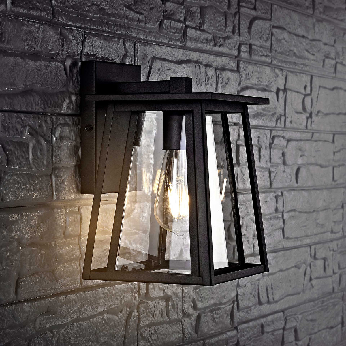 A black metal wall lantern casts warm light across textured masonry through its clear glass panels.