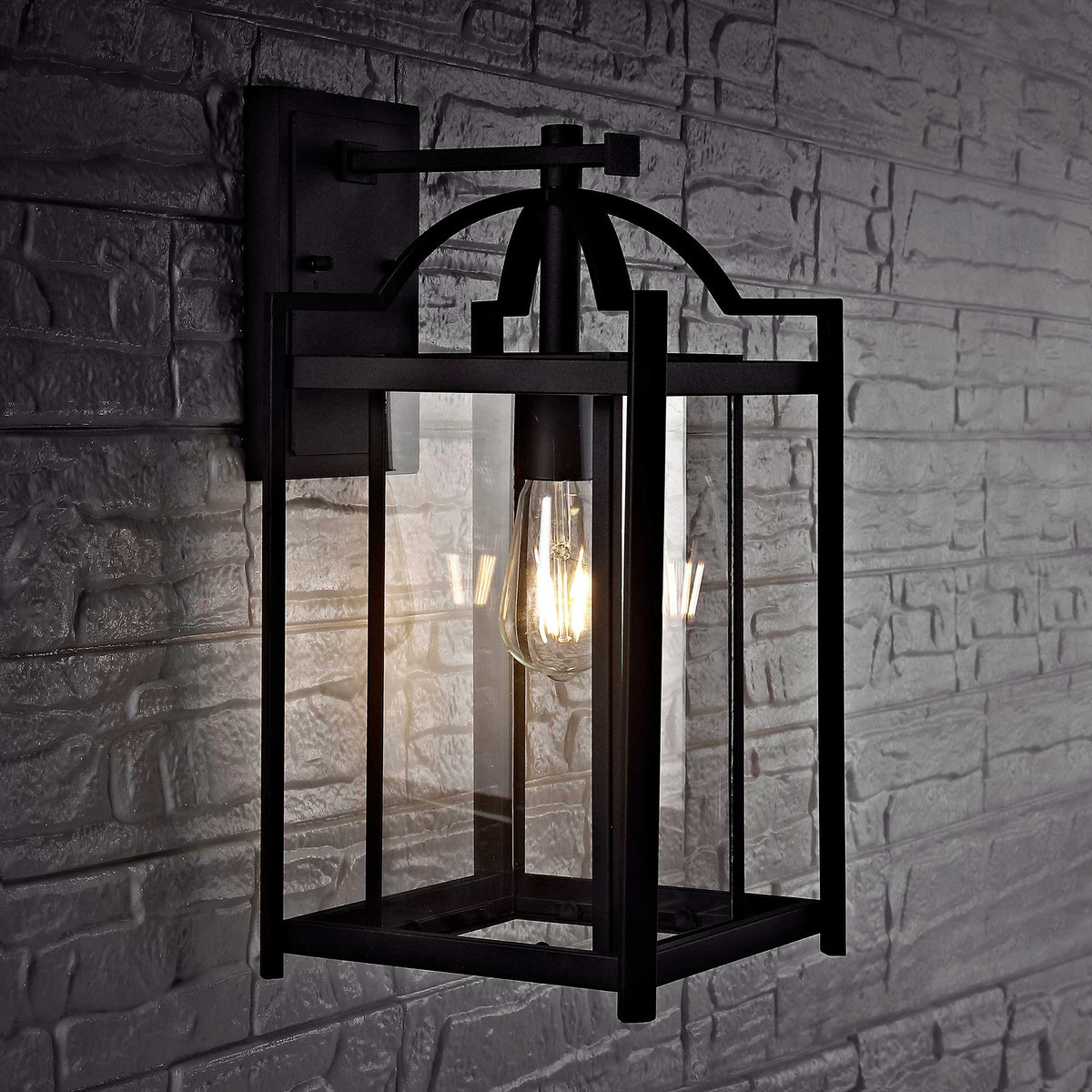 A glowing bulb shines through clear glass in a black wall lantern mounted on textured stone