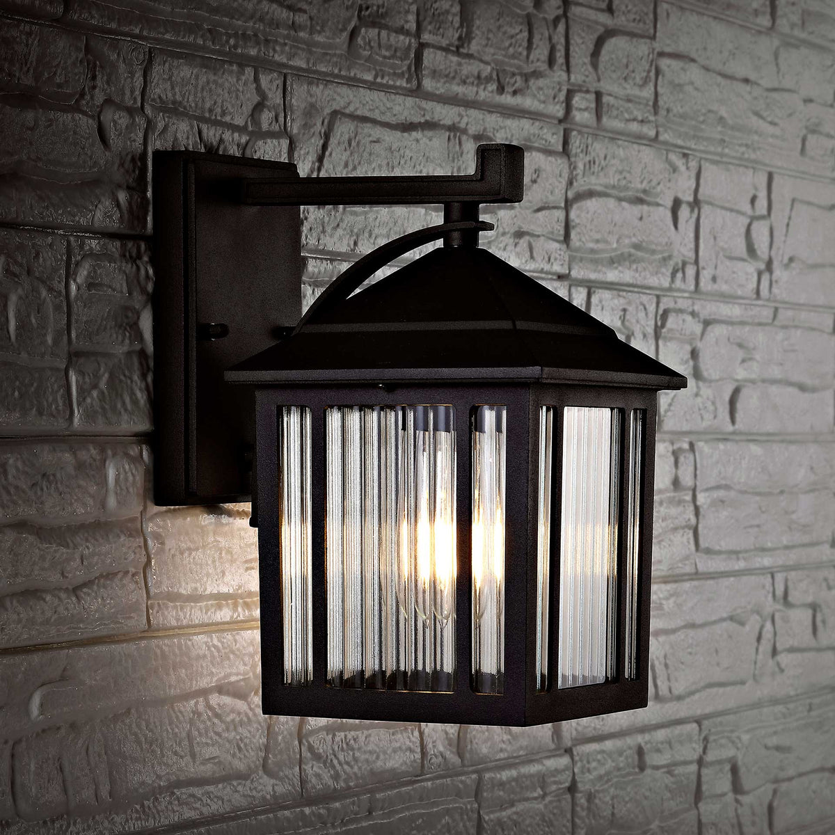 A black wall lantern casts a warm glow against textured stone through clear ribbed glass