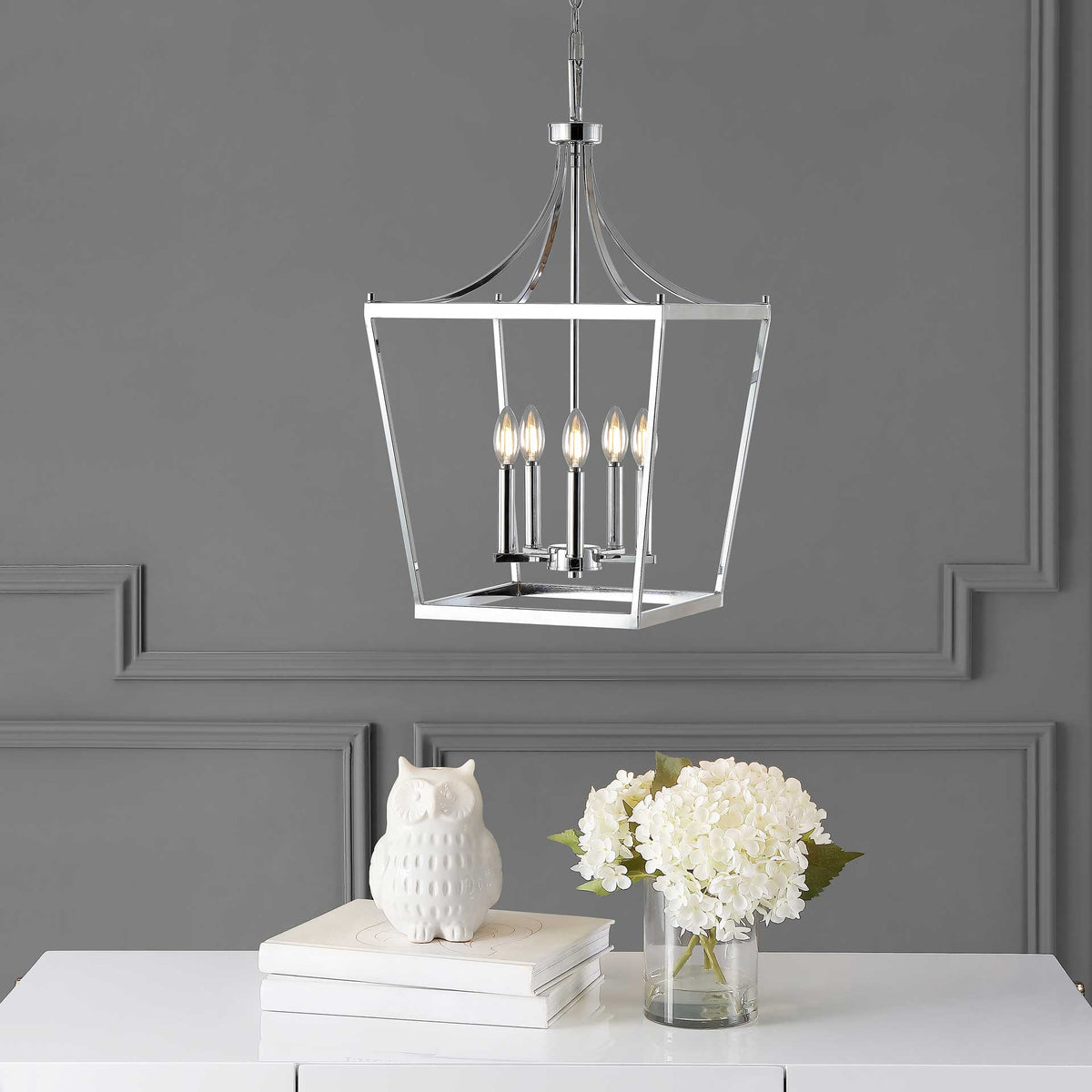 Warm candlelight shines through the pendant's open chrome frame above a white table.