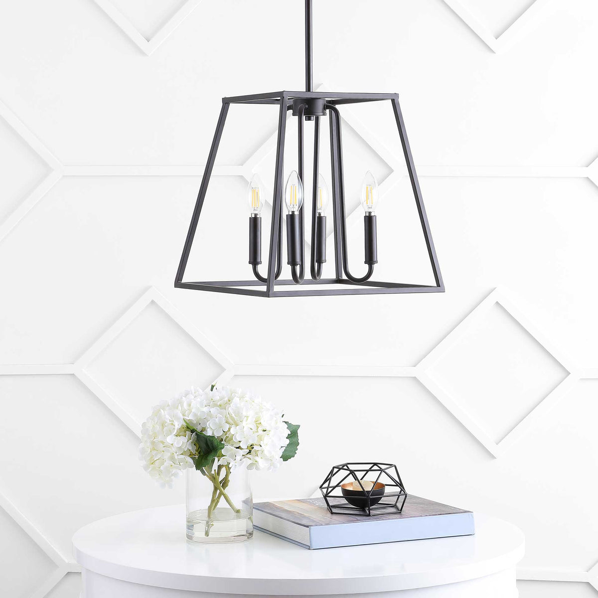 An angular bronze pendant with four candle-style bulbs hangs above a white table and flowers