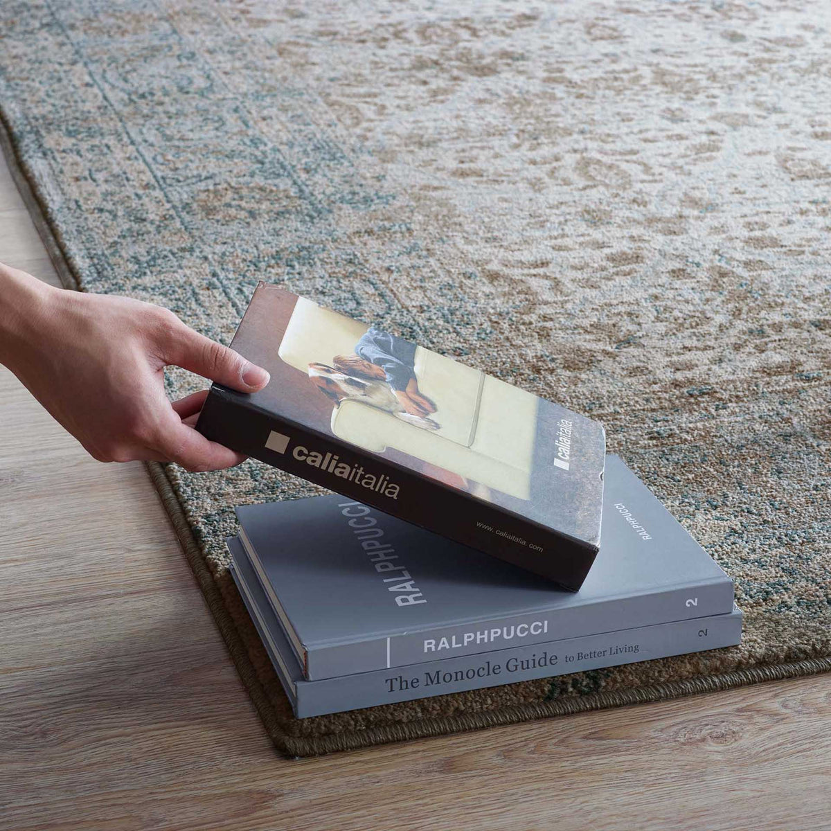 A low-pile floral rug lies flat on wood flooring beneath stacked books.