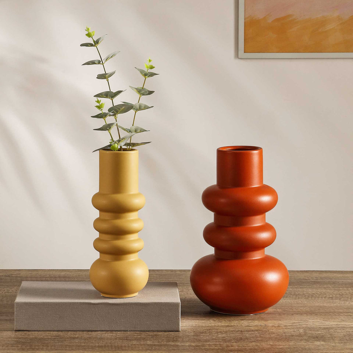 A pale yellow vase with leafy stems pairs with an orange vase for a colorful tabletop display