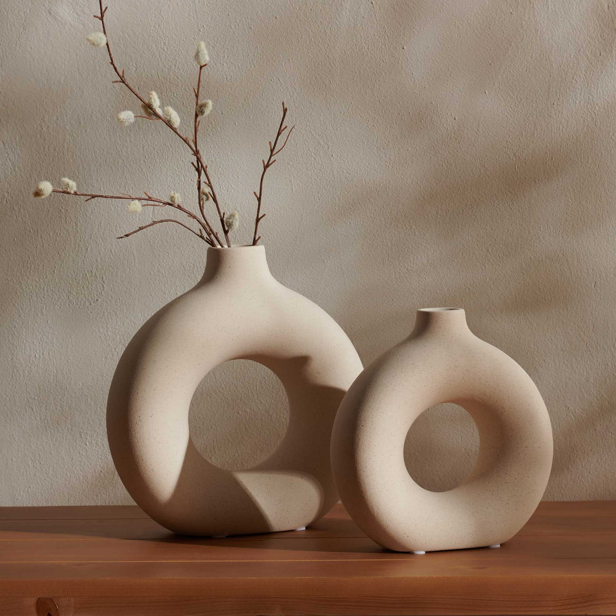 Two sculptural beige ceramic vases with open centers complement flowering branches on a tabletop.