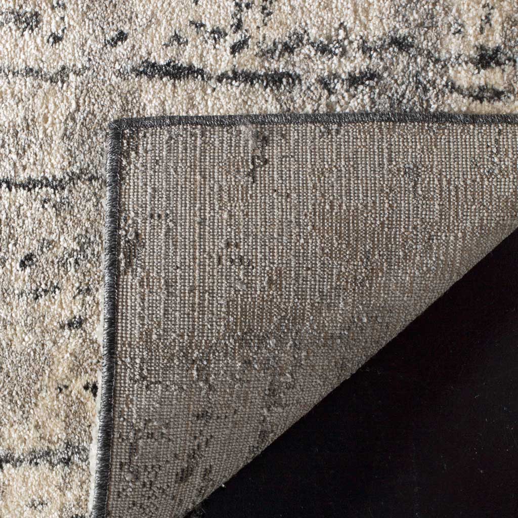 Folded corner reveals woven backing beneath light gray pile with scattered black markings