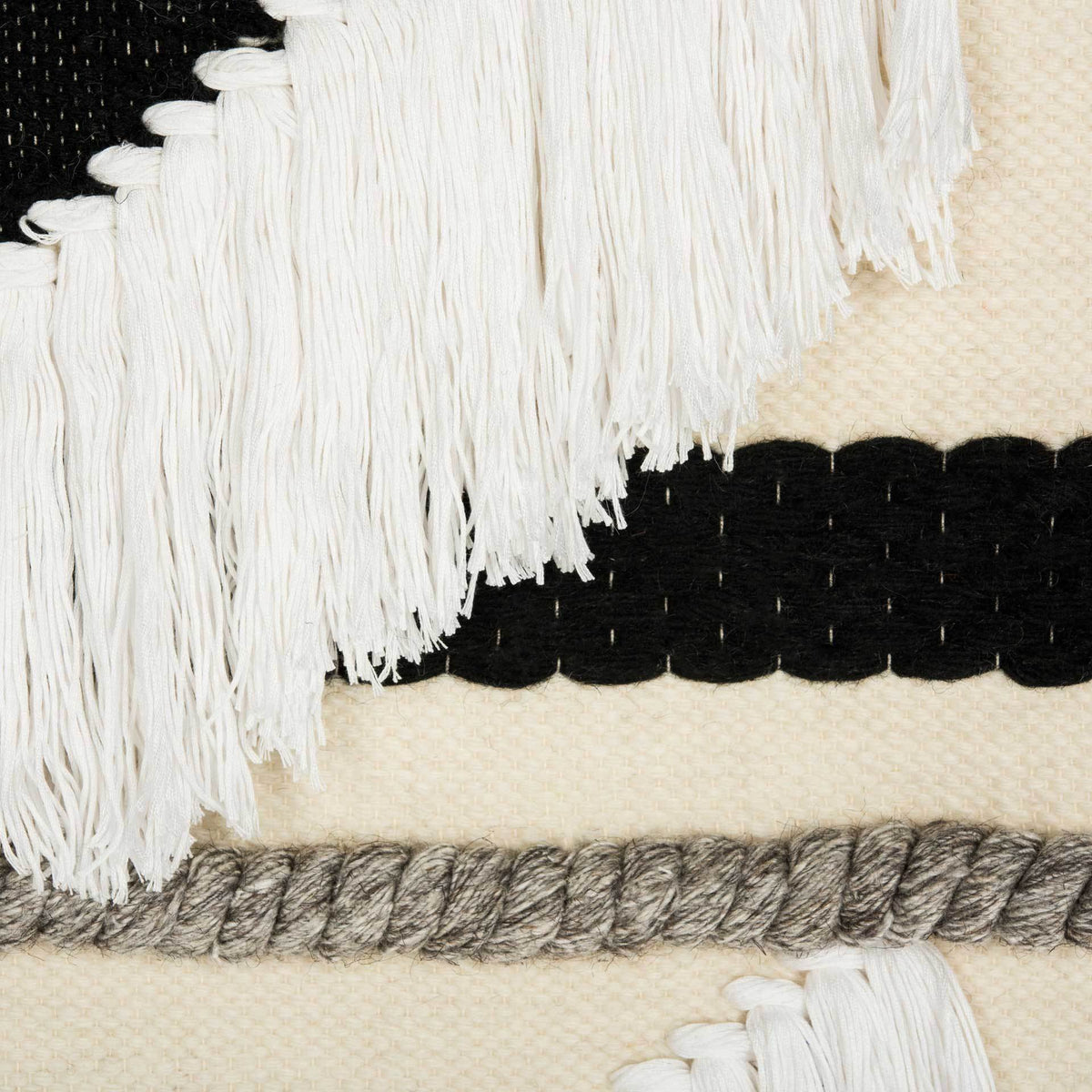 Cream tassels drape over bold black stripes and braided gray yarn in the tactile weave.