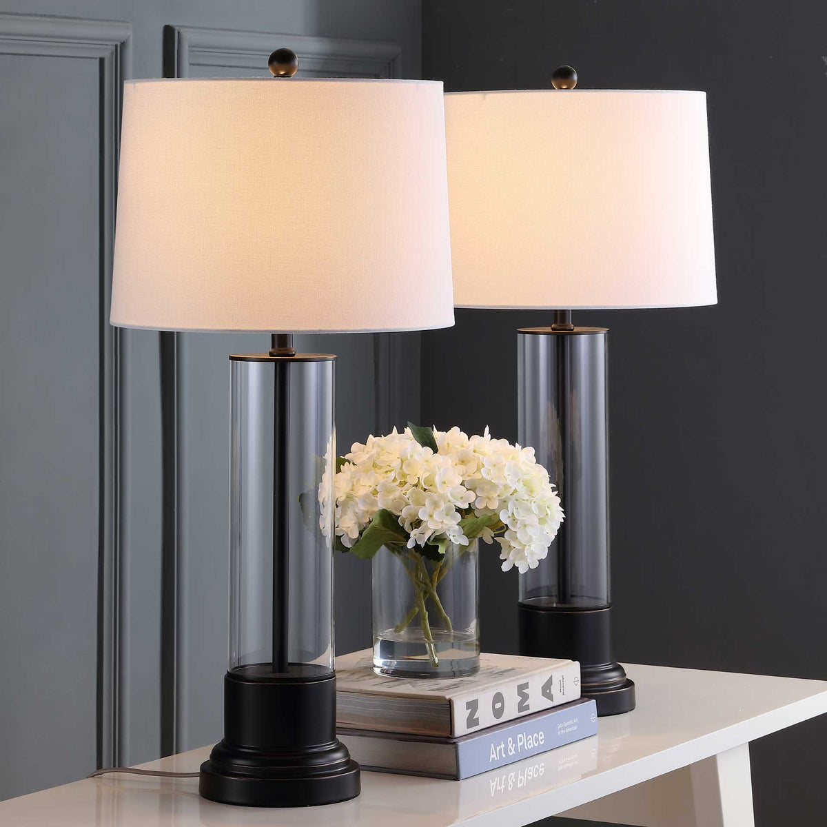 Two white fabric shades glow warmly above clear glass columns and black pedestal bases.