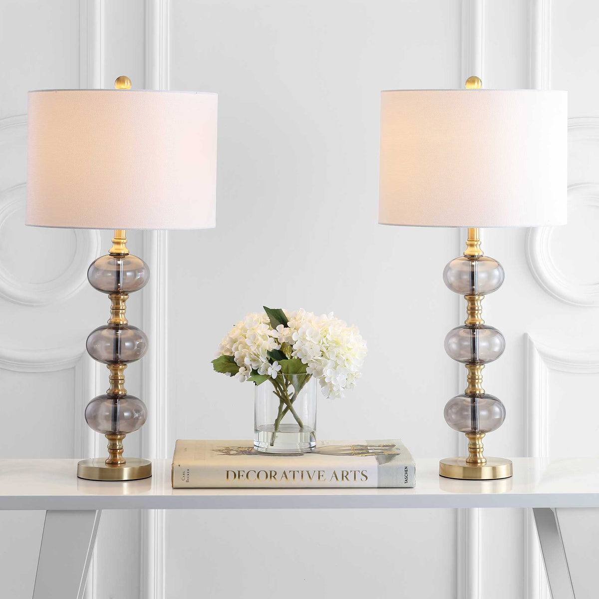 A glowing pair of lamps adds warm light with white drum shades, smoky glass orbs, and brass accents.