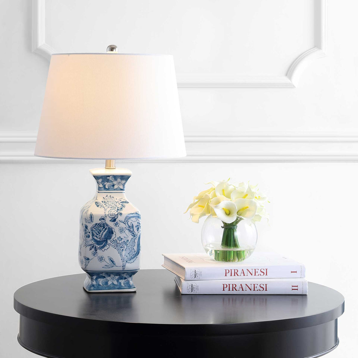A glowing white shade crowns a blue floral ceramic base beside a vase of flowers
