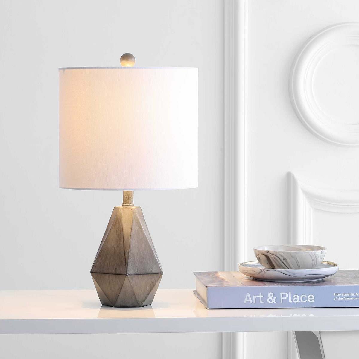 Warm light glows through the white drum shade above a sculptural dark gray base on a console table.