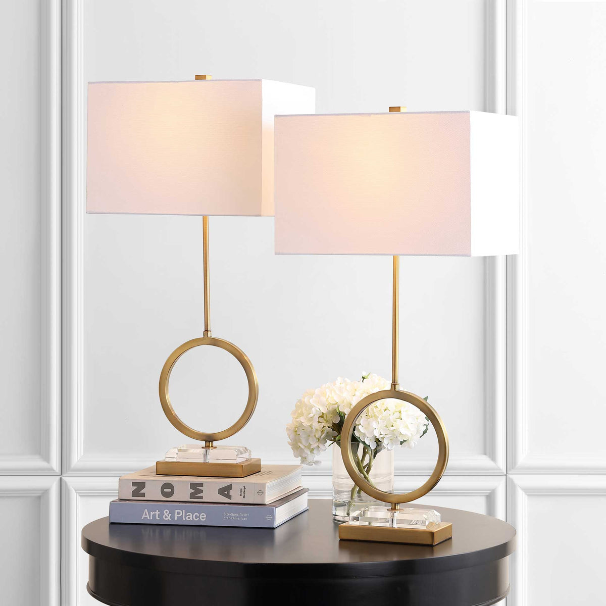 Warmly glowing shades crown two brass ring lamps with clear crystal bases beside flowers and books