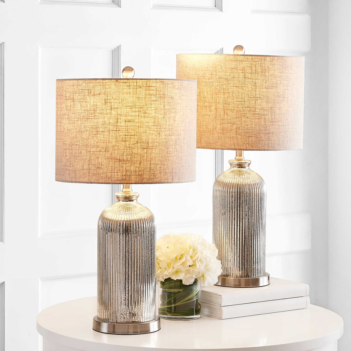 Warmly glowing drum shades brighten a pair of ribbed glass lamps beside white flowers.