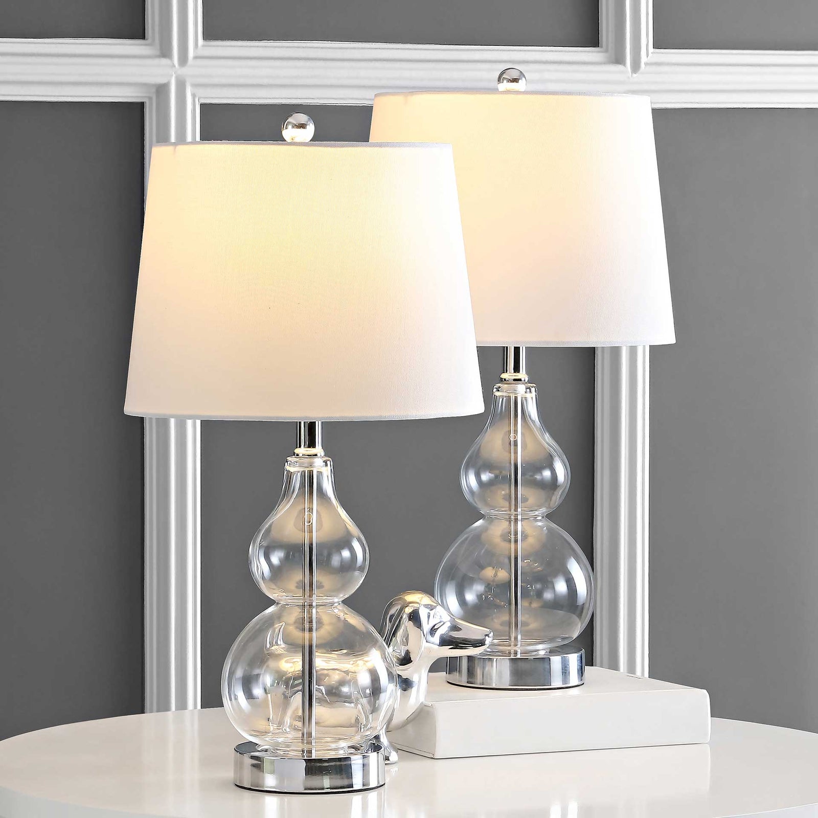 Two clear glass lamps glow warmly through white shades, with polished chrome accents below. || TBL4222A-SET2 || front_multi_on_room