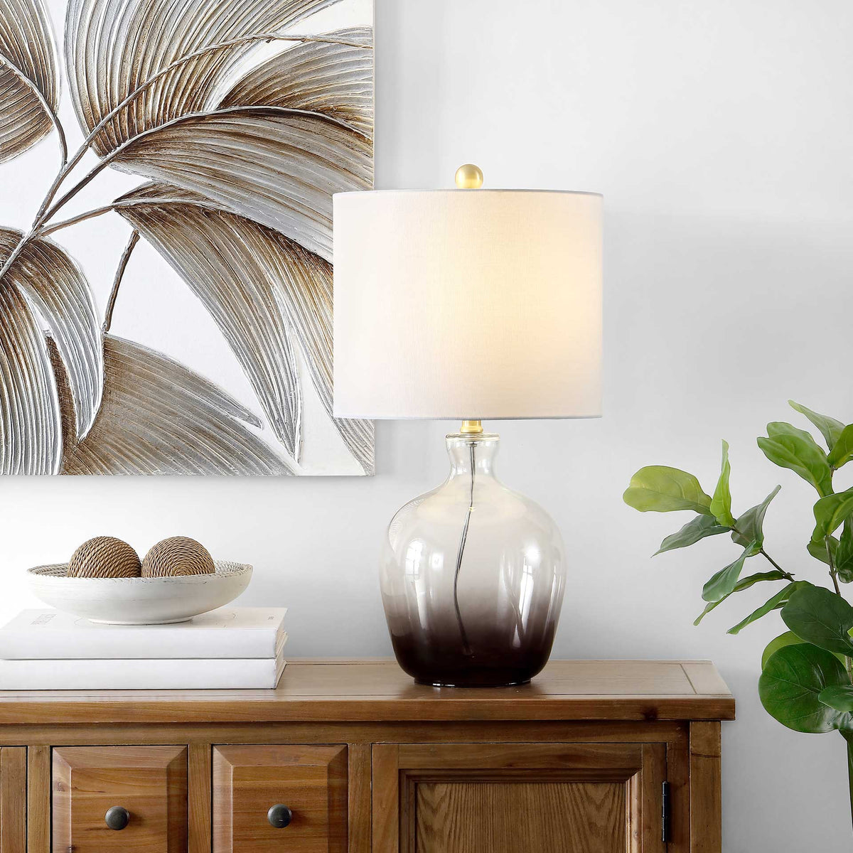 A warm glow shines through the white drum shade above a clear-to-smoky gray glass base